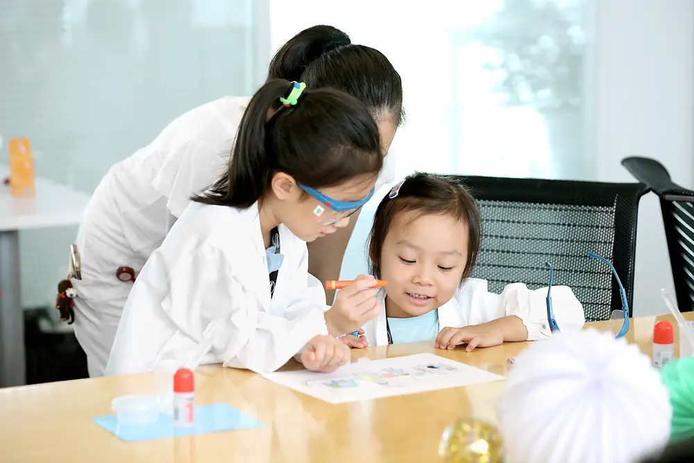 연구용 가운을 입고 있는 두 어린이와 여성이 테이블에서 그림을 색칠하고 있다