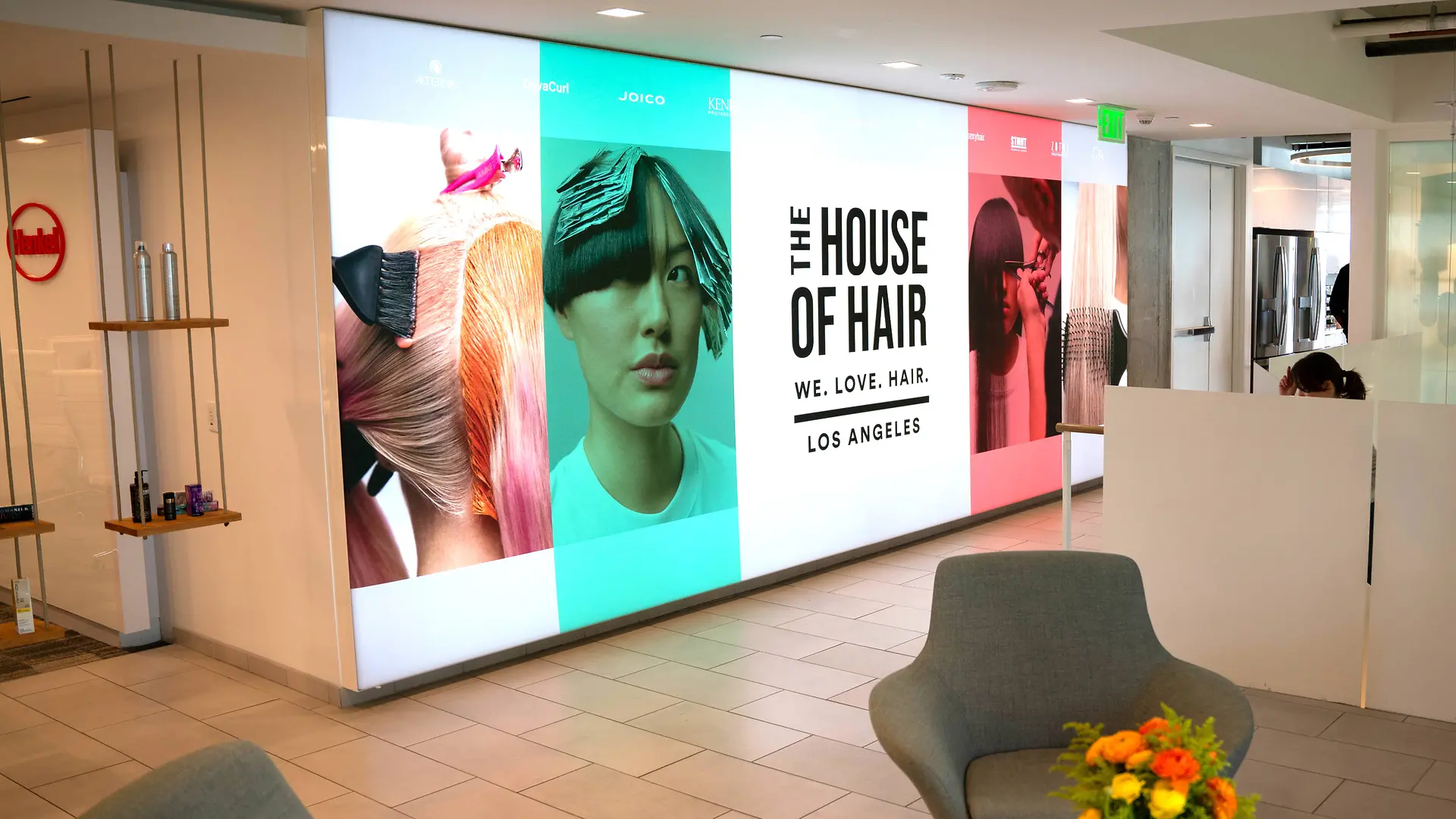 대형 컬러 헤어 그래픽과 “THE HOUSE OF HAIR – WE. LOVE. HAIR. – LOS ANGELES”라는 문구가 있는 밝은 로비 공간으로, 회색 라운지 체어와 꽃 장식 테이블이 배치된 미니멀한 디자인이 특징입니다.