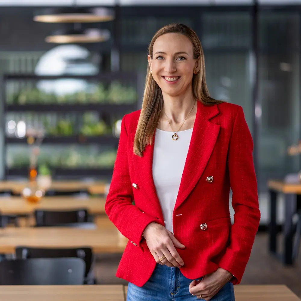 밝은 빨간색 재킷과 흰색 상의를 착용한 시몬 바겔-트라가 나무 테이블과 장식용 화병이 있는 현대적인 카페 스타일의 공간에 서 있는 모습.